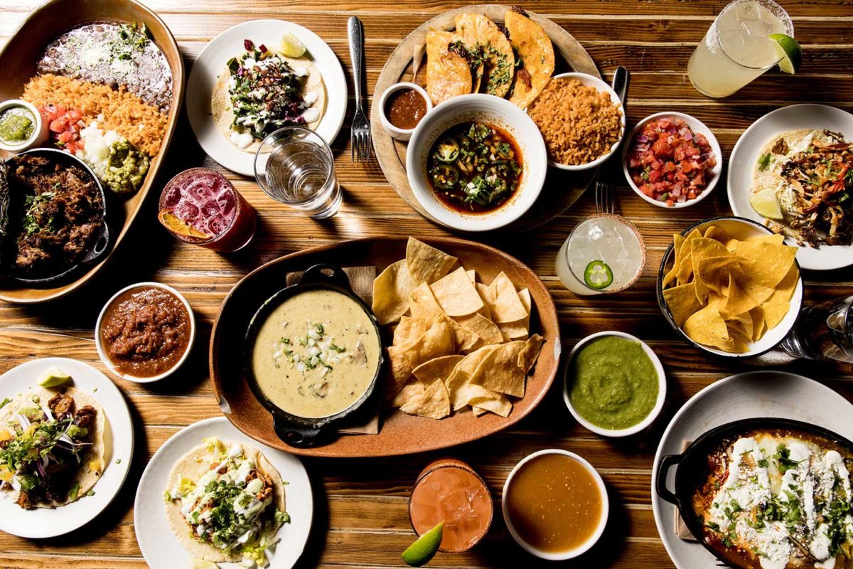 top down view of food including queso fundido, birria tacos, roasted street corn, and tacos.