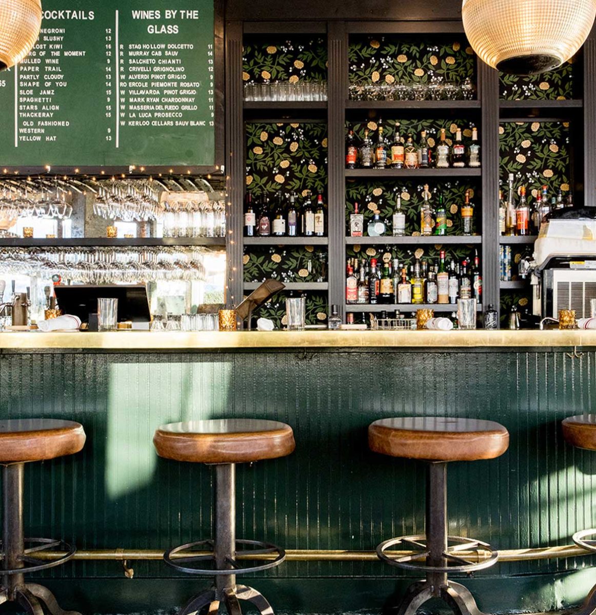 the bar at fiasco features a warm brass bar top, the backbar is lined with a modern floral wallpaper in shads of green and grass.